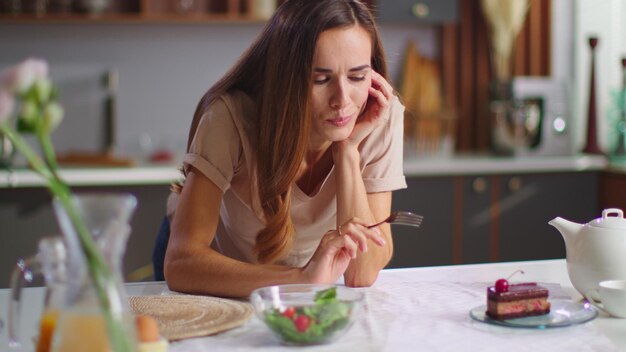 Photo femme réfléchie choisissant entre un gâteau et une salade dans la cuisine fille pointant avec une fourchette sur un délicieux gâteau et une salade saine sur la table femme faisant le choix entre une salade fraîche et un gâteau à la maison