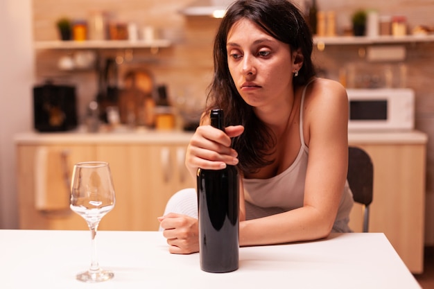 Femme réfléchie regardant un verre de vin assis sur la chaise. Personne malheureuse souffrant de migraine, de dépression, de maladie et d'anxiété se sentant épuisée par des symptômes de vertiges ayant des problèmes d'alcoolisme