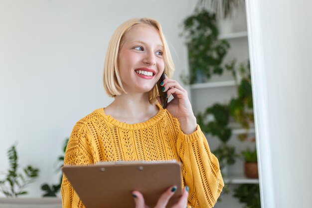 Femme réfléchie tenant du papier tout en parlant au téléphone mobile. La femelle regarde par la fenêtre. Elle est à la maison.