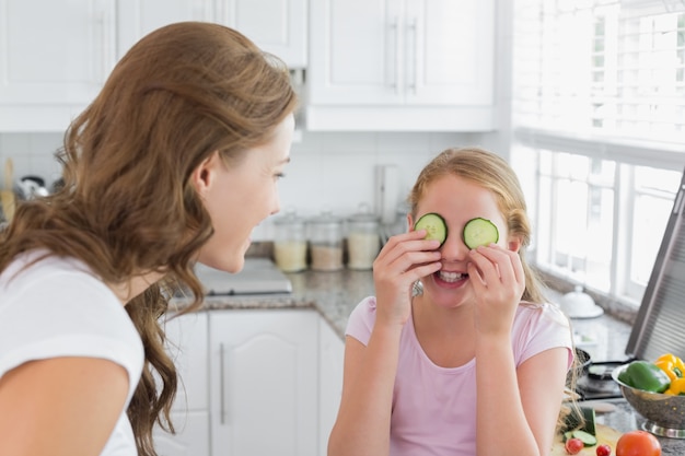 Femme regardant la fille garder les tranches de concombre sur les yeux dans la cuisine