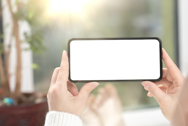 Une femme regarde du contenu vidéo ou joue à un jeu sur un téléphone portable ou un smartphone mobile avec une maquette d'écran blanc vierge à la maison