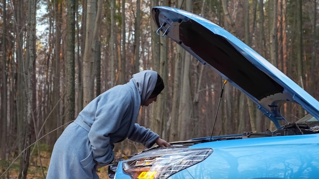 Une femme regarde sous le capot d'une voiture cassée debout sur une route forestière