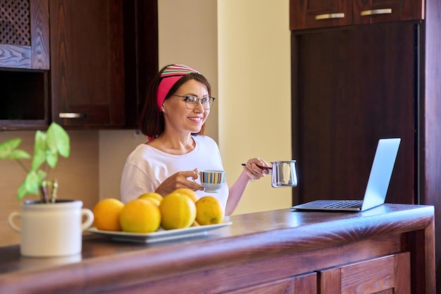 Femme relaxante souriante avec café du matin avec cezve turc et ordinateur portable dans la cuisine à la maison