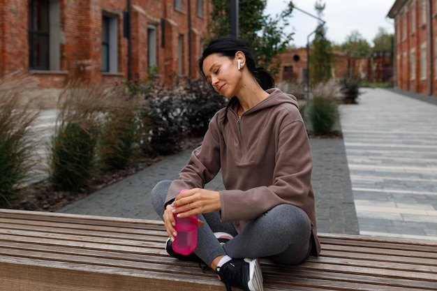 Une femme de remise en forme dans un casque sans fil et une bouteille de sport dans les mains est assise sur un banc et écoute de la musique après une séance d'entraînement en plein air