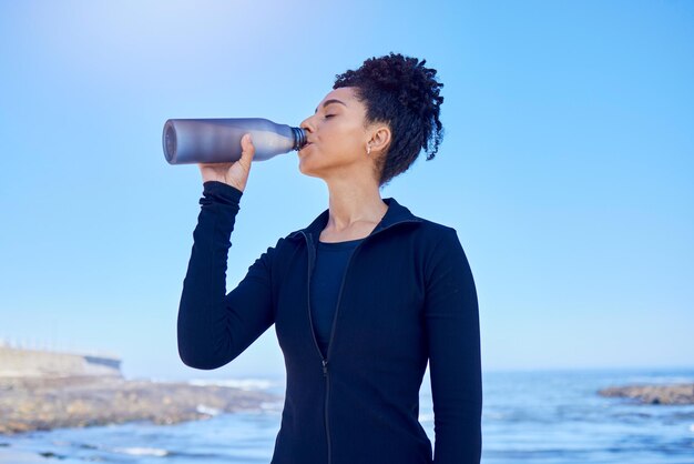 Femme de remise en forme et eau potable à la plage pour courir, santé ou cardio matinal sur un ciel bleu au bord d'un océan. Faire de l'hydratation et une coureuse assoiffée avec un liquide de sport après un entraînement ou un entraînement en mer.