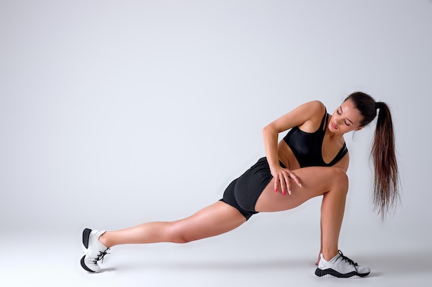 Femme de remise en forme faisant des exercices de fentes pour l'entraînement musculaire des jambes, une athlète active fait un exercice de fente avant vers l'avant d'une jambe, sur fond de studio blanc, vue latérale