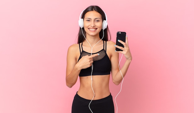 Femme de remise en forme hispanique à l'air excité et surpris en pointant sur le côté avec des écouteurs et un téléphone