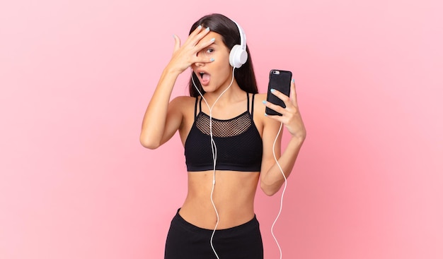 Femme de remise en forme hispanique ayant l'air choquée, effrayée ou terrifiée, couvrant le visage avec la main avec des écouteurs et un téléphone