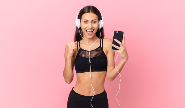 Femme de remise en forme hispanique se sentant choquée, riant et célébrant le succès avec des écouteurs et un téléphone