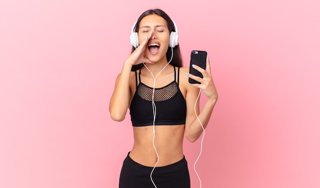 Femme de remise en forme hispanique se sentant heureuse, donnant un grand cri avec les mains à côté de la bouche avec des écouteurs et un téléphone