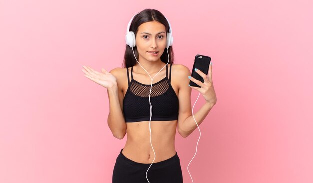 Femme de remise en forme hispanique se sentant perplexe et confuse et doutant avec des écouteurs et un téléphone