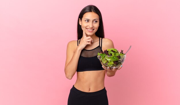 Femme de remise en forme hispanique souriant joyeusement et rêvant ou doutant et tenant une salade