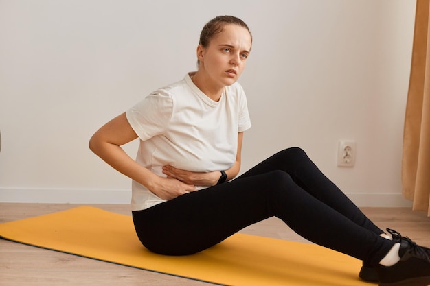 Femme de remise en forme portant un T-shirt blanc et des leggins noirs ayant des douleurs pendant l'entraînement à domicile. Malheureuse femme assise sur le tapis de yoga, ressentant des douleurs à l'estomac. Soins de santé et exercice.