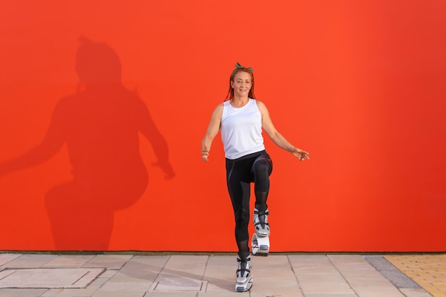 Femme de remise en forme rousse latina en vêtements de sport faisant une routine d'entraînement portant des bottes Kangoo Jumps sur fond orange. Mouvement actif, action, forme et bien-être