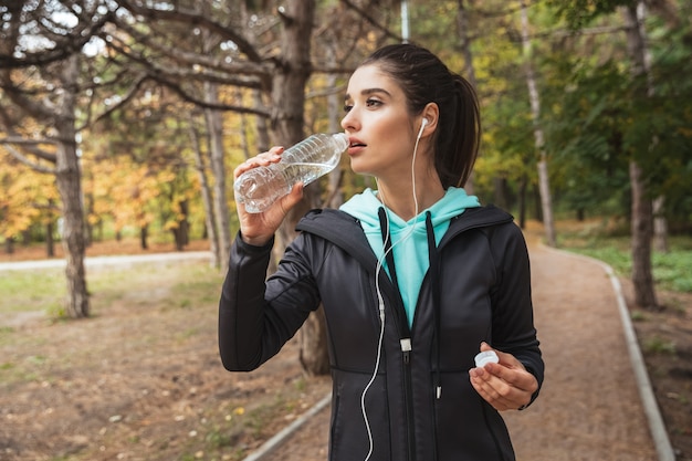 Femme de remise en forme souriante, écouter de la musique avec des écouteurs, tenant une bouteille d'eau en se tenant debout dans le parc