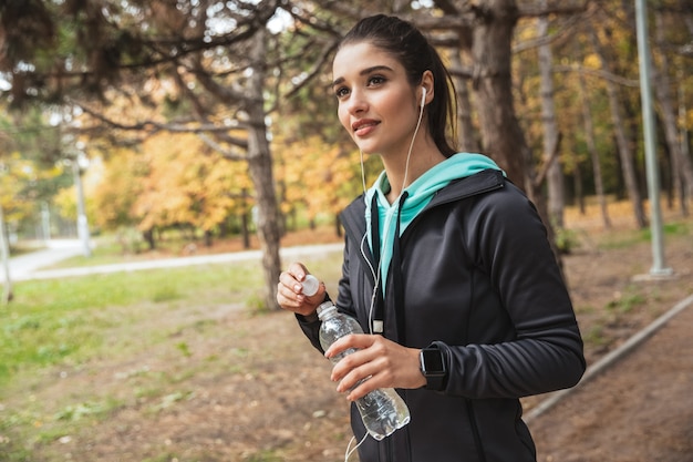 Femme De Remise En Forme Souriante, écouter De La Musique Avec Des écouteurs, Tenant Une Bouteille D'eau En Se Tenant Debout Dans Le Parc