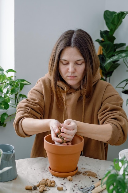 Une femme remplissant un pot de fleurs en argile avec de l'argile expansée pour le drainage se préparant à rempoter une plante d'intérieur