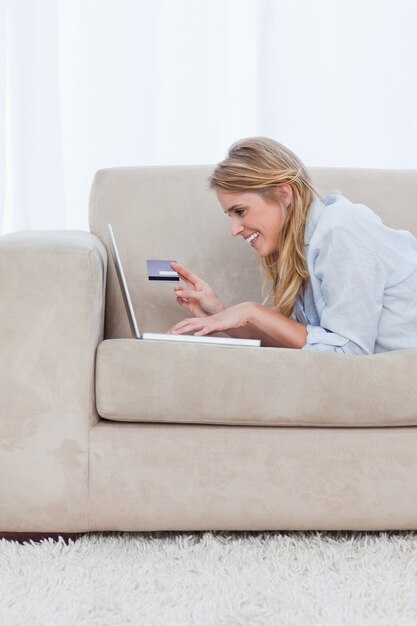 Une femme reposant sur ses coudes tient une carte bancaire et tape sur son ordinateur portable