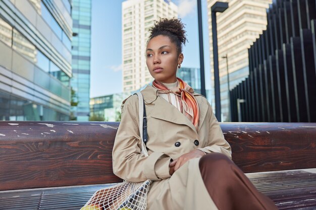 la femme repose sur un banc en bois dans la grande ville contre les gratte-ciel porte un imperméable regarde loin porte un sac net