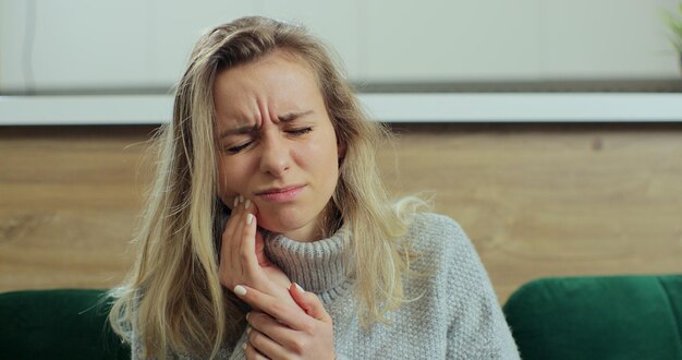 Femme ressentant une forte douleur dans la dent tout en restant à la maison. Famale blonde souffrant de maux de dents alors qu'elle était assise sur un canapé. Concept d'hygiène bucco-dentaire et de soins dentaires.