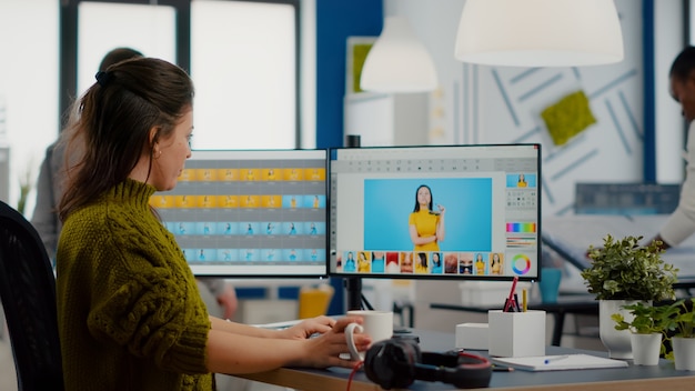 Femme retoucheuse regardant la caméra souriante assise dans une agence de création multimédia retouchant des photos de clients sur PC avec deux écrans