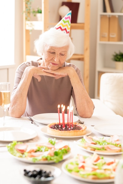 Femme à la retraite âgée à la recherche de bougies allumées sur un gâteau fait maison alors qu'il était assis par table d'anniversaire servi