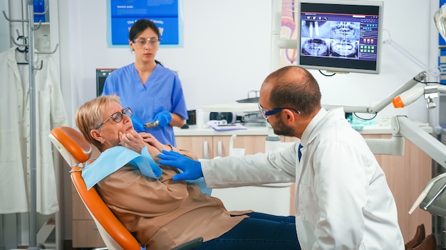 Femme à la retraite présentant des symptômes de douleur aux gencives tenant la main sur la joue tout en parlant avec le médecin. Patient âgé expliquant le problème dentaire au médecin indiquant la bouche pendant que l'infirmière prépare des outils stériles.