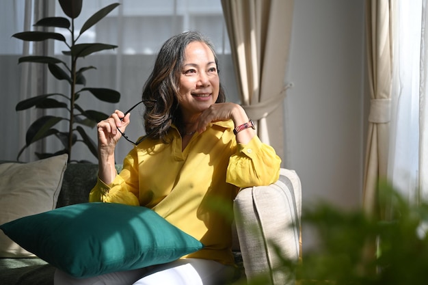 Femme à la retraite tenant des lunettes et regardant par la fenêtre tout en se reposant sur un canapé à la maison