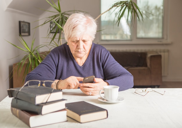 Une femme à la retraite utilise un téléphone portable dans une cuisine confortable. Grand-mère fait une pause-café en cuisine