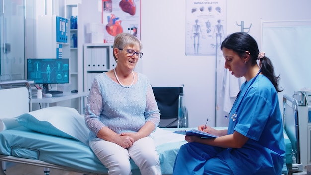 Femme retraitée senior ayant une discussion avec une infirmière dans un hôpital moderne, assise sur son lit et répondant aux questions avant la consultation de rendez-vous en médecine médicale. Salle de diagnostic en cli moderne privé