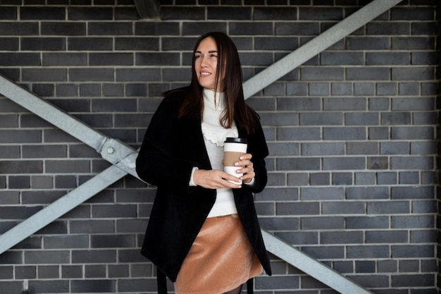 Femme réussie adulte avec une tasse de café et un sourire sur son visage contre un mur de briques