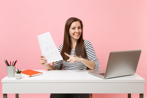 Femme réussie pointant l'index sur des documents papier, travaillant sur un projet alors qu'elle était assise au bureau avec un ordinateur portable