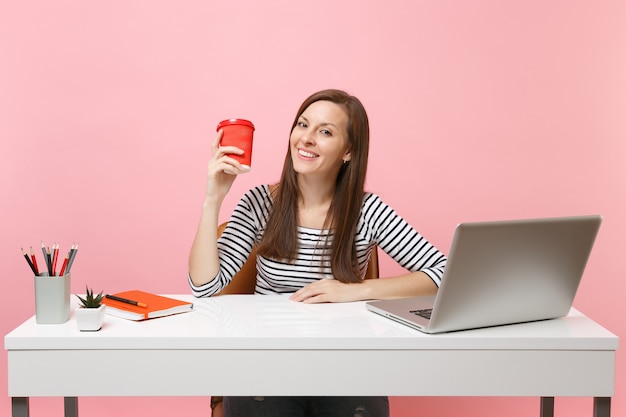 Femme réussie tenant une tasse de café ou de thé travaillant sur un projet assis au bureau avec un ordinateur portable