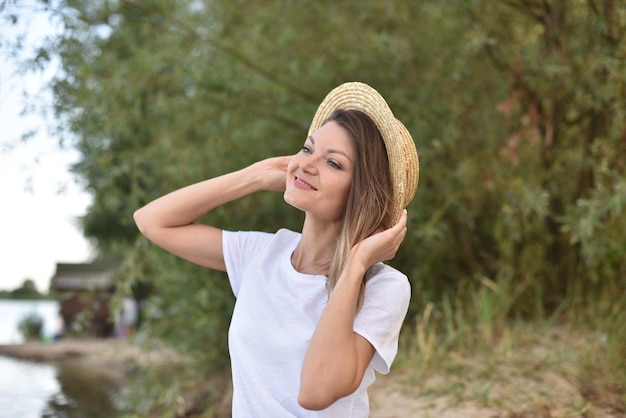 femme rêvant en t-shirt blanc tient un chapeau de paille avec les mains et regarde le ciel bleu assis sur le rivage