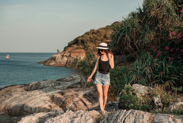 Femme rêveuse sur une grosse pierre à la plage tropicale portant un chapeau en osier. Concept de voyage de vacances d'été