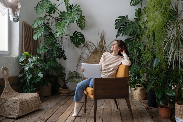 Une femme rêveuse se repose après avoir terminé le travail sur un ordinateur portable, regarde dans la fenêtre assise sur une chaise dans un jardin confortable