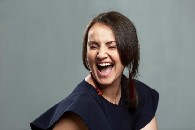 Femme riant émotionnellement avec la bouche ouverte. Belle brune en robe bleue et boucles d'oreilles élégantes. Joie, bonheur et positivité. Fond gris.