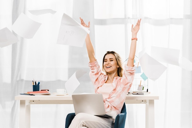 Femme riante avec un ordinateur portable jetant des documents sur le lieu de travail