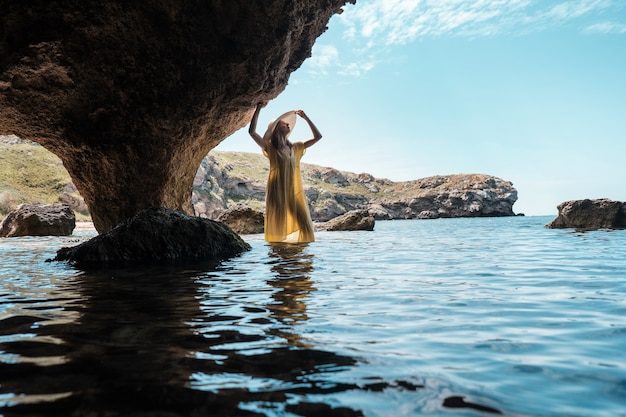 Une femme en robe blanche et chapeau noir debout sur les rochers au bord de la mer