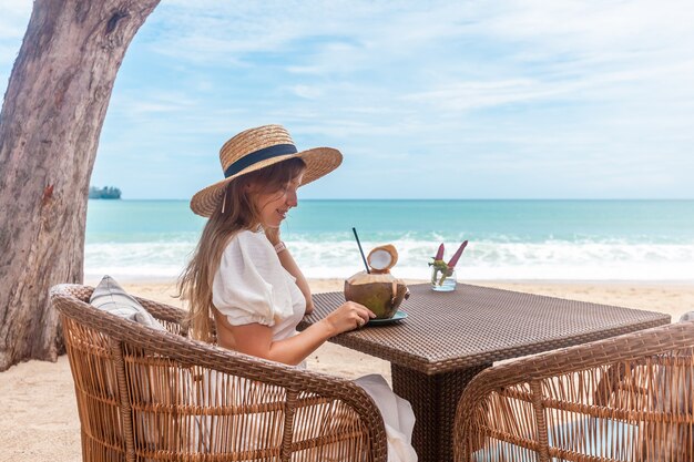 Femme en robe blanche et chapeau de paille assis sur une table dans un café de plage en plein air