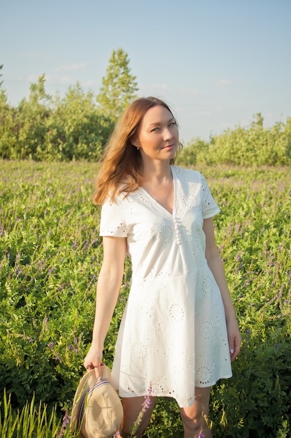 femme en robe blanche sur un pré. Elle se tient avec un chapeau dans ses mains.