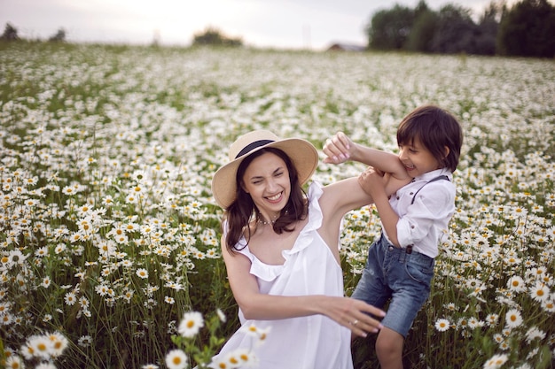 Femme en robe blanche avec son fils un garçon de 5 ans se trouve dans un champ de camomille au coucher du soleil en été