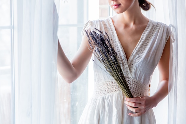 Femme en robe blanche tenant la lavande dans les mains et rester près de la fenêtre. Image dans un style high key