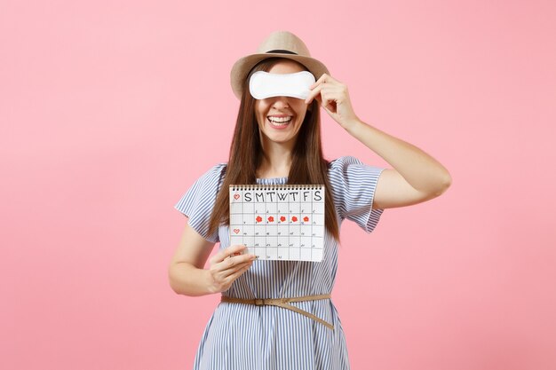 Femme en robe bleue, chapeau tenant une serviette hygiénique, yeux de couverture, calendrier des périodes féminines pour vérifier les jours de menstruation isolés sur fond rose. Concept médical, sanitaire, gynécologique. Espace de copie