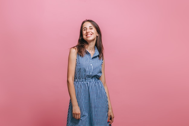 Une femme en robe bleue se dresse sur un mur rose et sourit