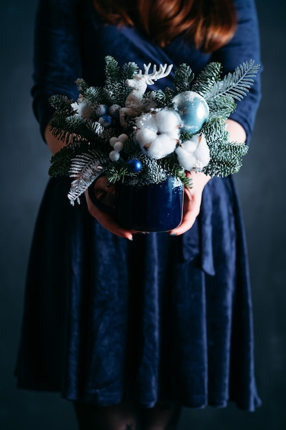 femme en robe bleue tenant un arrangement de sapin à la main avec des cerfs, du coton, de la fausse neige.