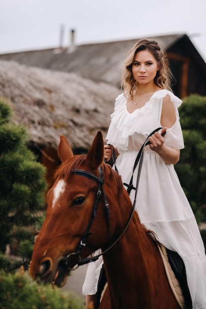 Une femme en robe d'été blanche à cheval près d'une ferme