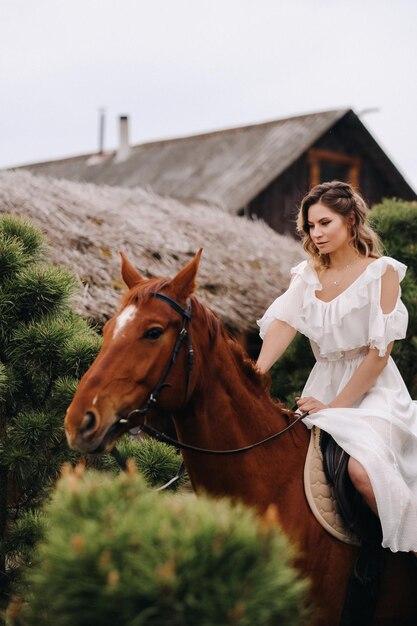 Une femme en robe d'été blanche à cheval près d'une ferme