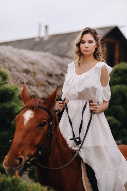 Une femme en robe d'été blanche à cheval près d'une ferme