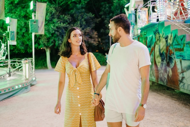 Une femme en robe jaune au décolleté plongeant et un homme barbu marchent en se tenant la main entre des manèges. Un couple d'amoureux en rendez-vous à la foire de Valence.